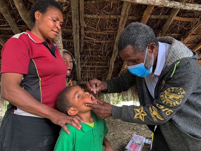 A health worker holds a child's chin and dips their head back to administer a polio vaccine. The child's mother rest her hands on his shoulders.